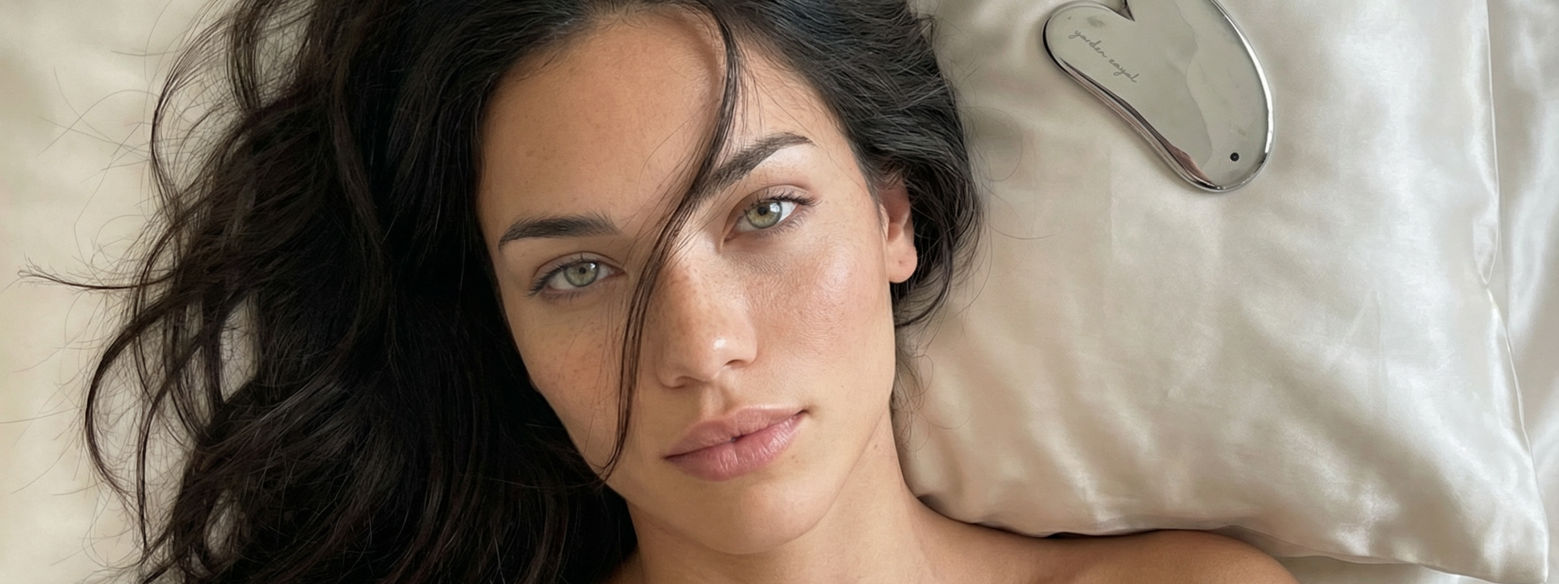 Frau mit braunen Haaren, hellblauen Augen und softem Glow liegt auf dem Bett und hat eine Strähne im Gesicht. Ihr Kopf liegt auf dem Kissen und auf dem Kissen ist ein Terahertz Gua Sha zu sehen.