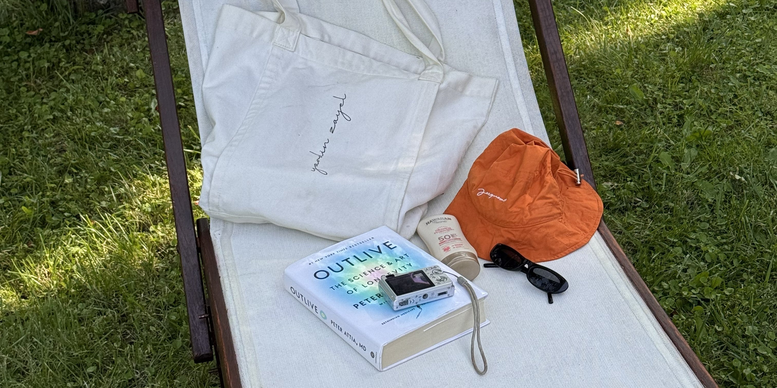 Liegematte mit der Flowtote von yardin zayel, dem Buch OUTLIVE, einer Kamera, Sonnencreme, Sonnenbrille und Sonnenhut darauf