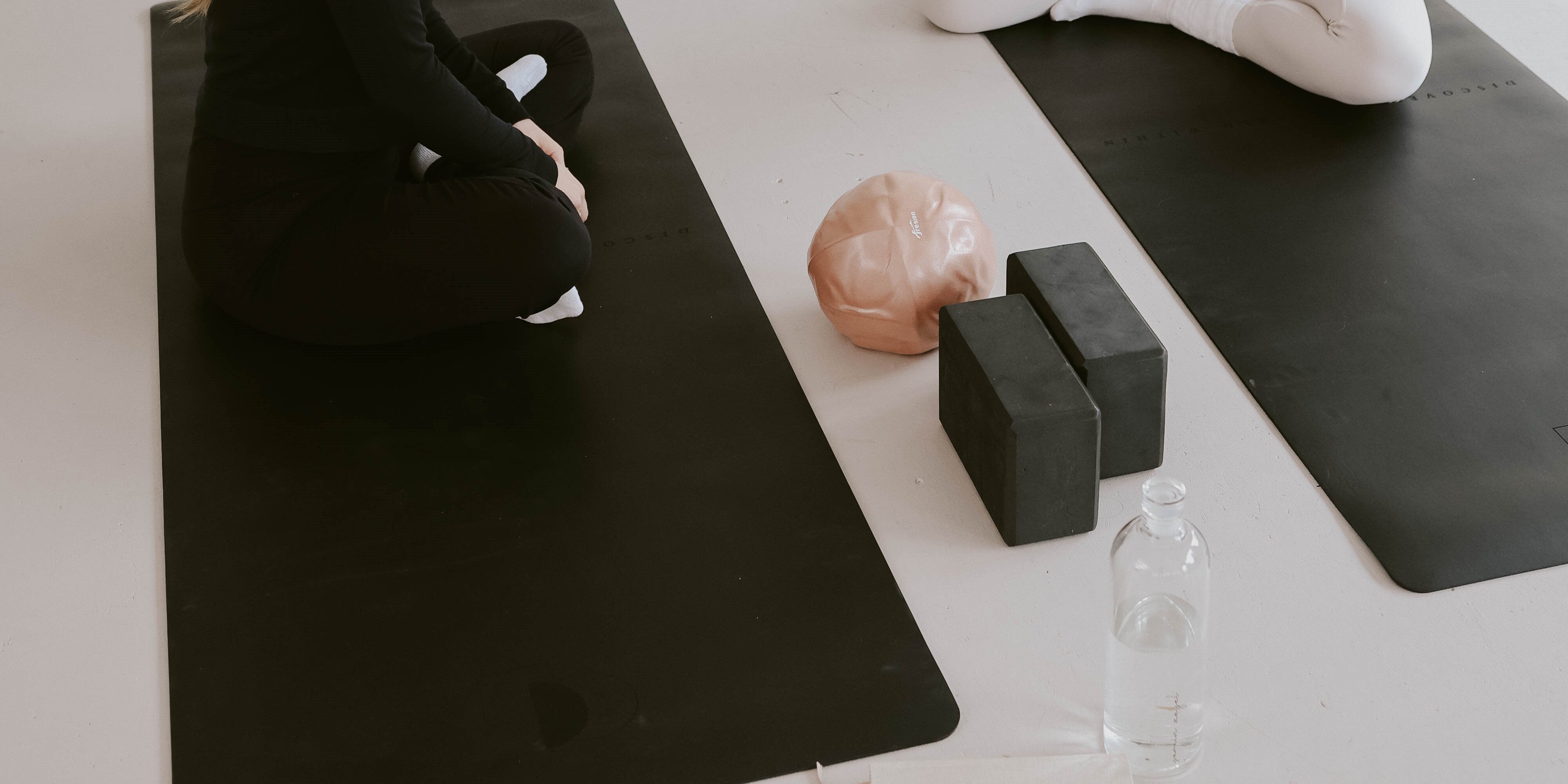 Zwei sitzende Frauen auf Yogamatten, Yogablöcke, ein rosa Pilatesball und eine Glasflasche voll mit Wasser