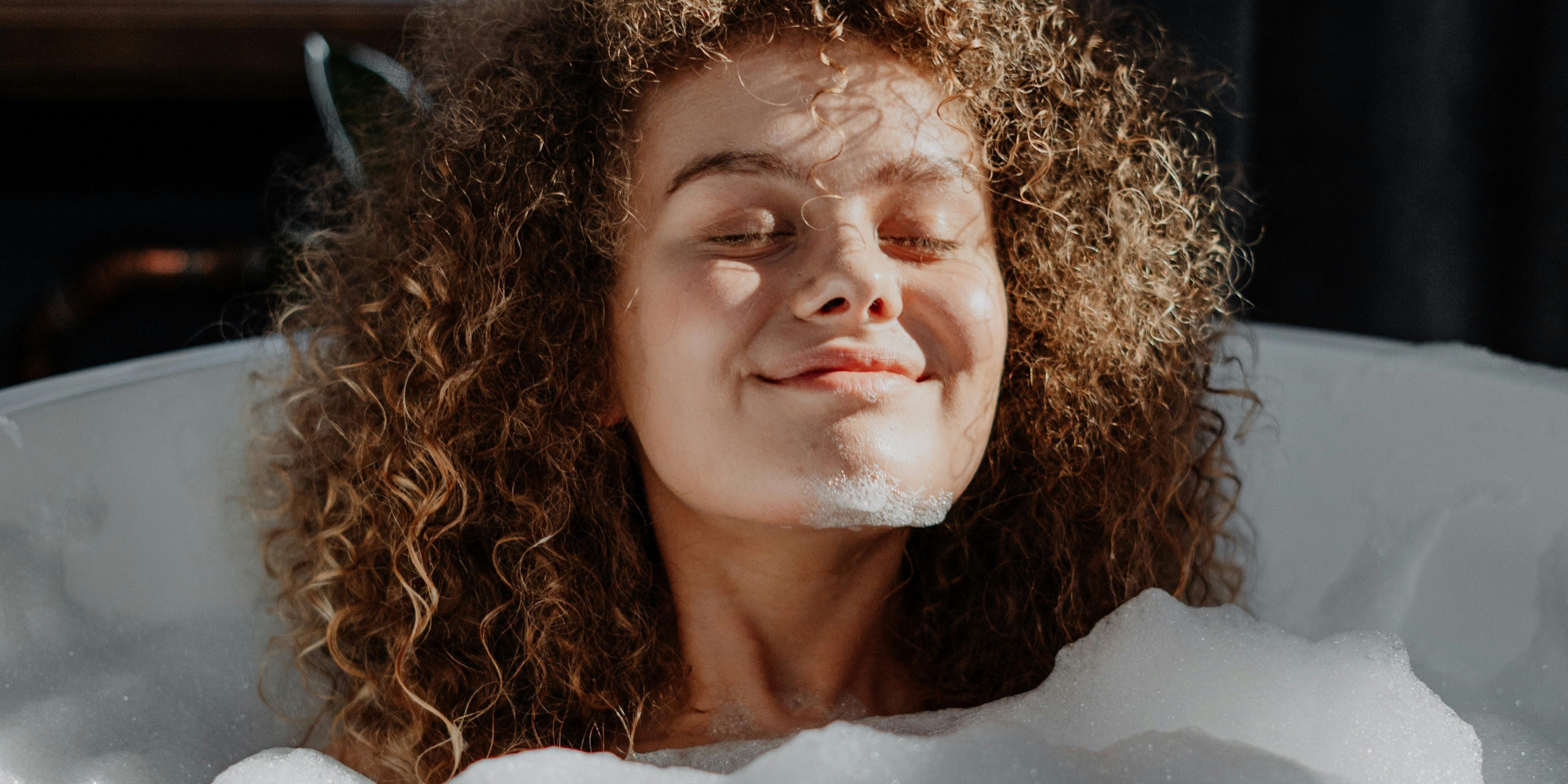 Braunhaarige Frau mit Locken die in einer Badewanne voll Schaum sitzt und Lächelt während sie von der Sonne angestrahlt wird