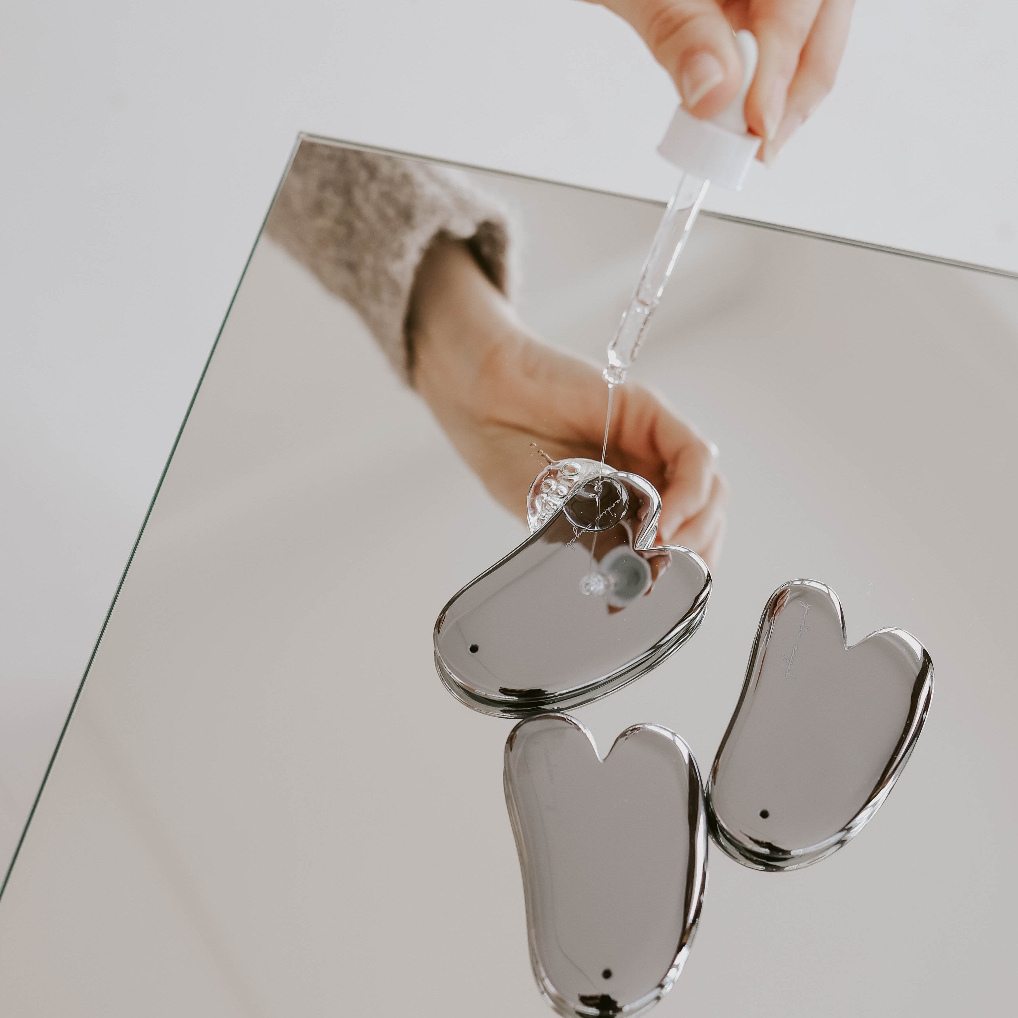Es liegen drei Teraglow Gua Sha aus Terhertz auf einem spiegelnden Beistelltisch. Eine Hand lässt von oben ein durchsichtiges Serum auf den Terglow tropfen.
