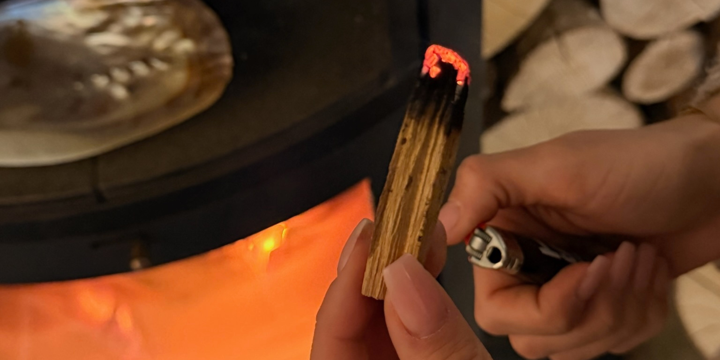 Hand entzündet ein Palo-Santo-Holzstück mit Feuerzeug; die Spitze glüht, warmes Kaminfeuer und Muschel im Hintergrund, ruhige Selfcare-Atmosphäre.