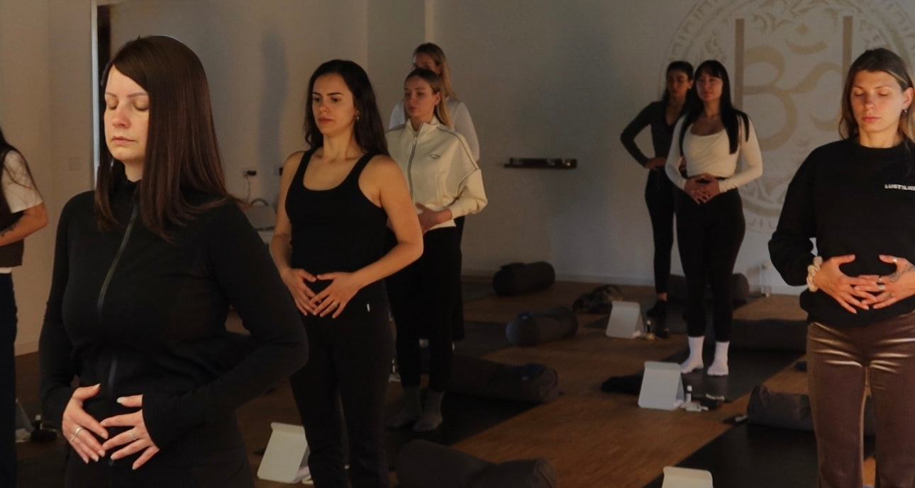 sieben Frauen stehen in einem Yogastudio, haben die Augen geschlossen, heben sich die Hände auf dem Bauch und sind gerade mitten in einer Breathwork Session.