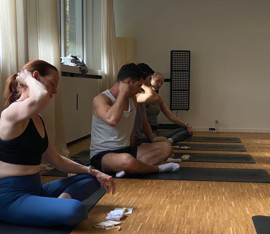 zwei Männer und zwei Frauen sitzen auf schwarzen Yogamatten von yardin zayel und haben ein Gua Sha in der Hand, während sie ihren Hals damit massieren. Im Hintergrund sieht man ein Redlight Panel, das für Redlight Therapy genutzt wird.