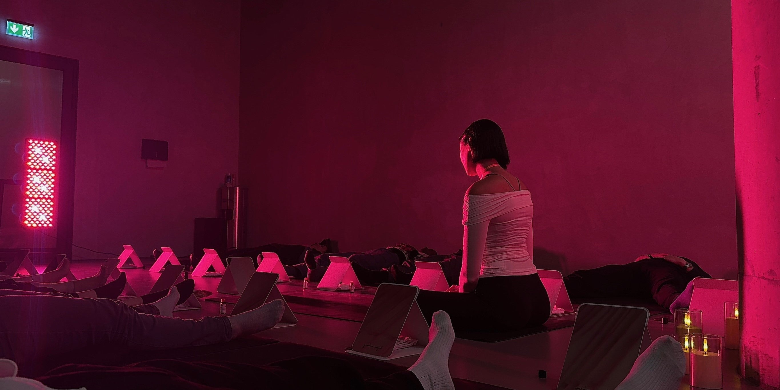 Teilnehmer einer Breathwork Session die auf Yogamatten liegen und eine Frau die sitzt und die Gruppe anleitet, begleitet von Redlight