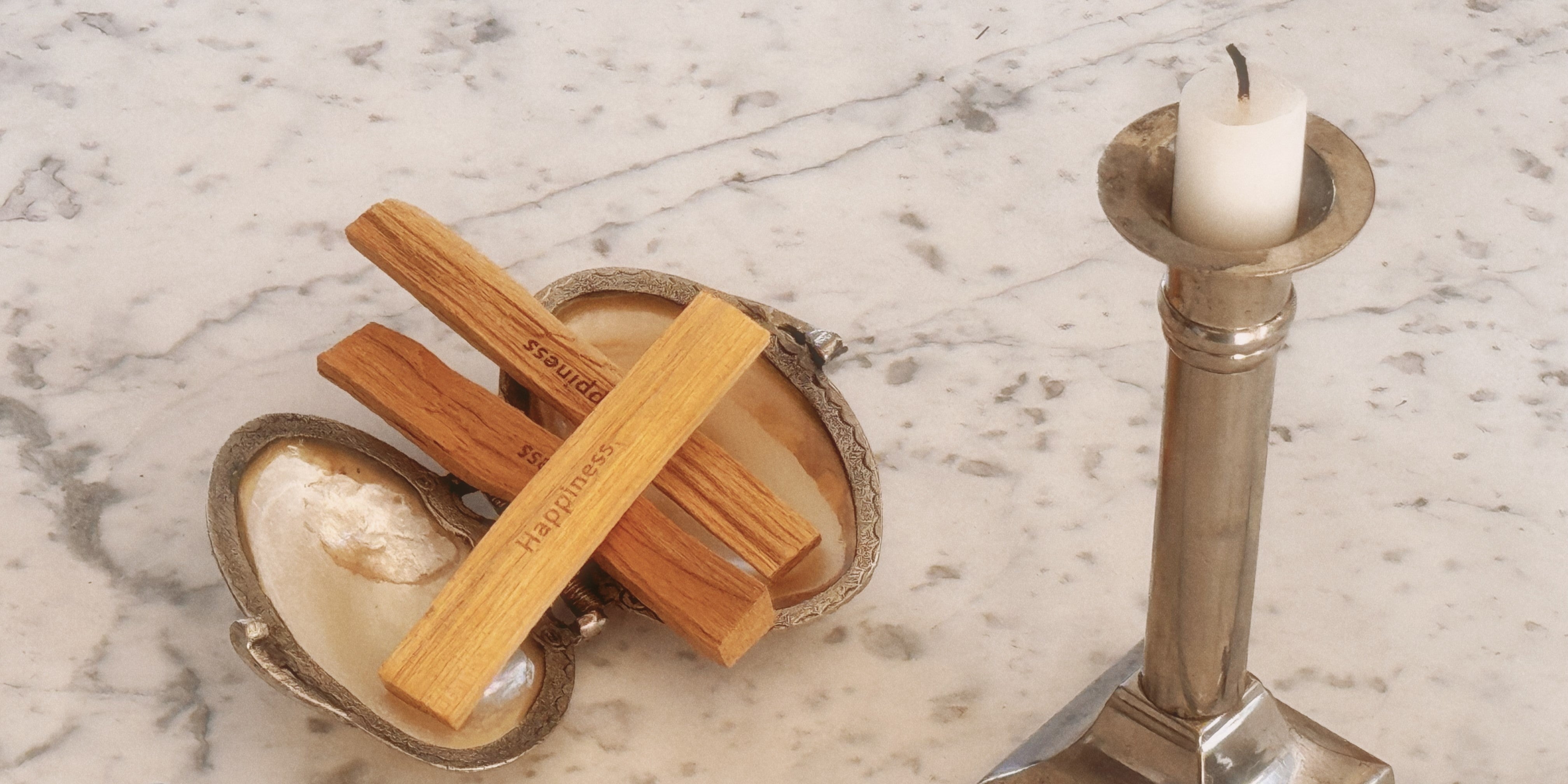 Palo Santo Sticks mit der Gravur Happiness in einer Muschel mit einer Kerzen in einem Silberkerzenständer auf einem Marmortisch