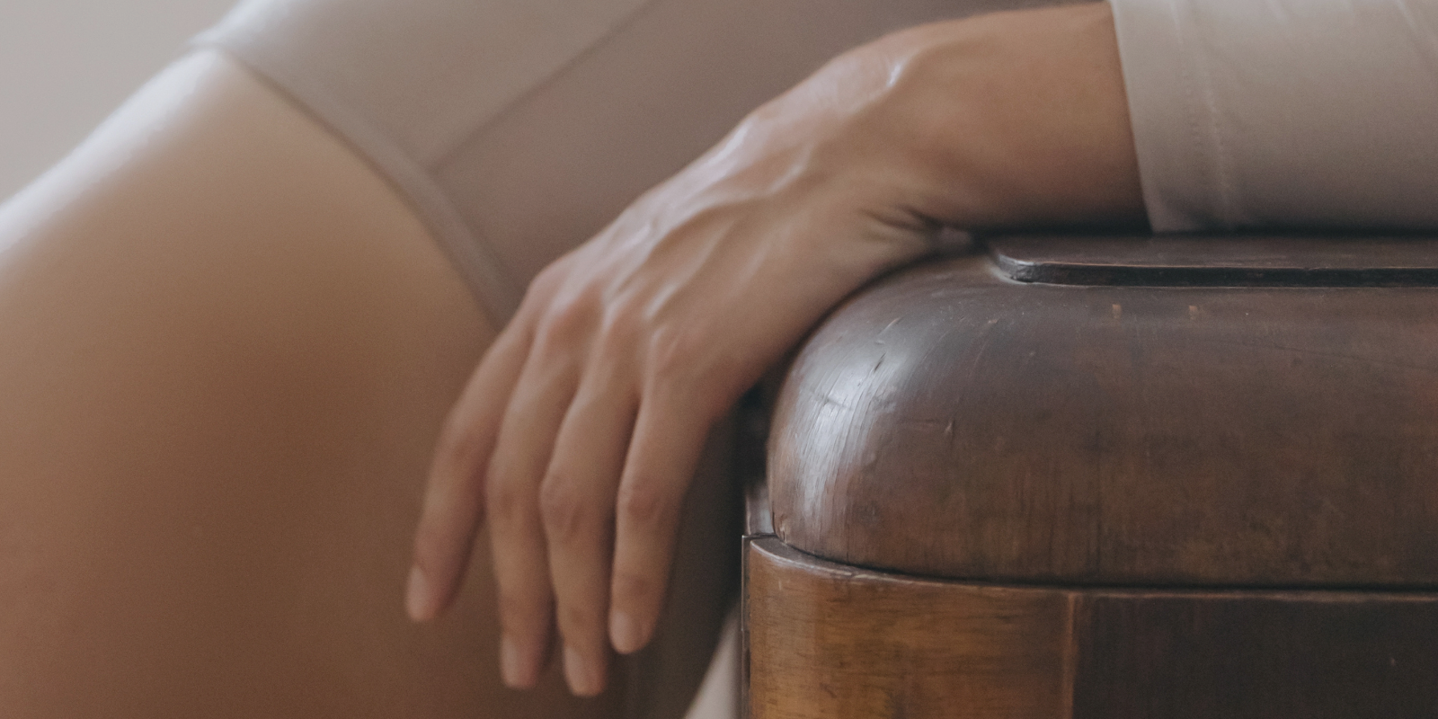 Eine weiblicher Körper, in einem beigen langärmeligen Body, welcher sich an einer Massivholz-Ablage anlehnt.