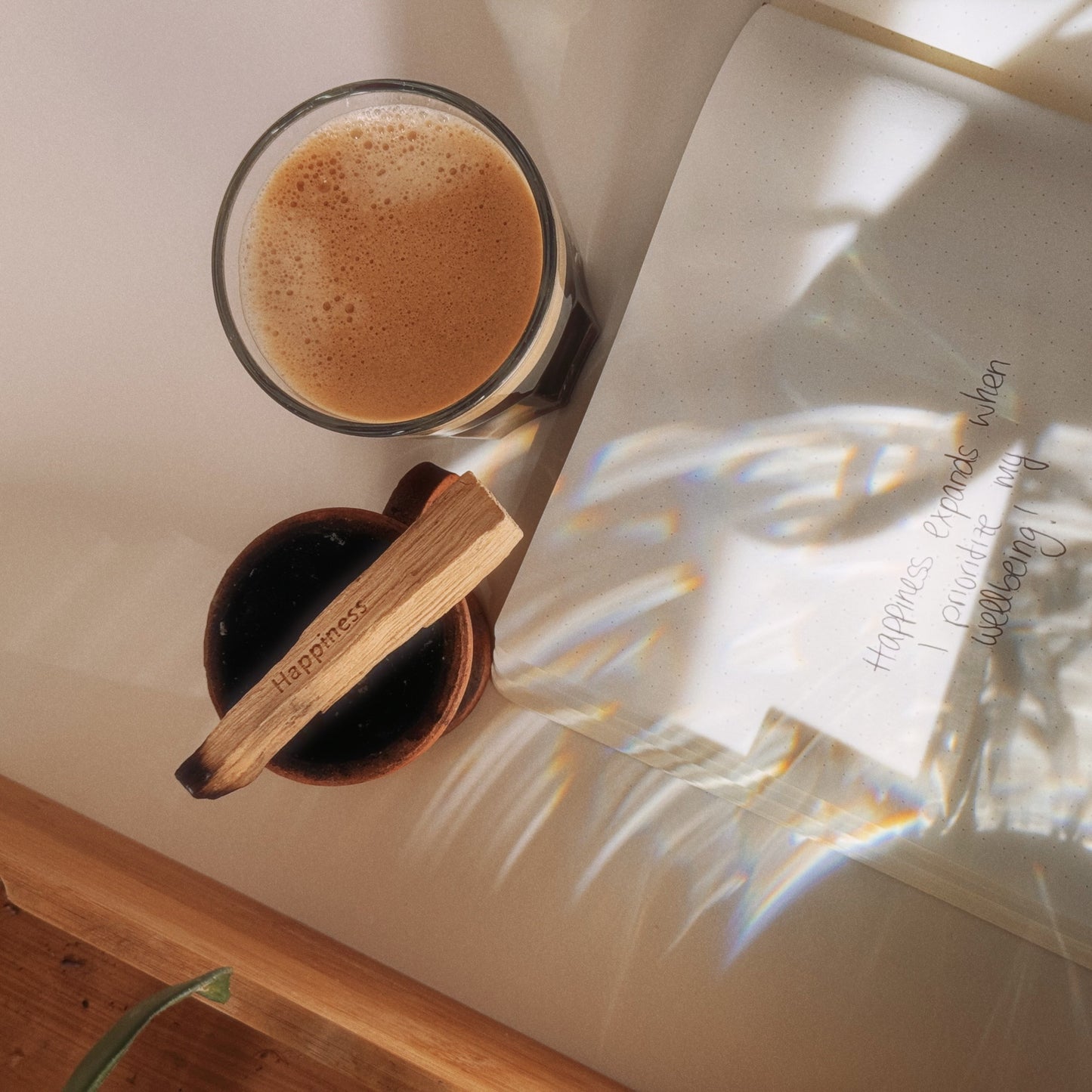 Harmonisches Bild mit Sonneneinflutung. Links oben ist ein angezündetes Palo Santo mit Mantra "Happiness". Es raucht nach oben. Rechts daneben ein Glas mit Kaffee. Zudem ist ein Journal zu sehen, mit der Affirmation "Happiness expands when I prioritize my wellbeing"
