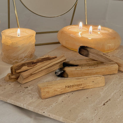 Nahaufnahme von Palo Santo Holz auf einem Steintablett, drei sind schon angekohlt, jeder hat eine andere Aufschrift: 'yardin zayel' 'Happiness' 'Love' 'Peace' 'Success', dahinter sind Teelichthalter aus Marmor mit brennenden Teelichtern und ein Tischspiegel.