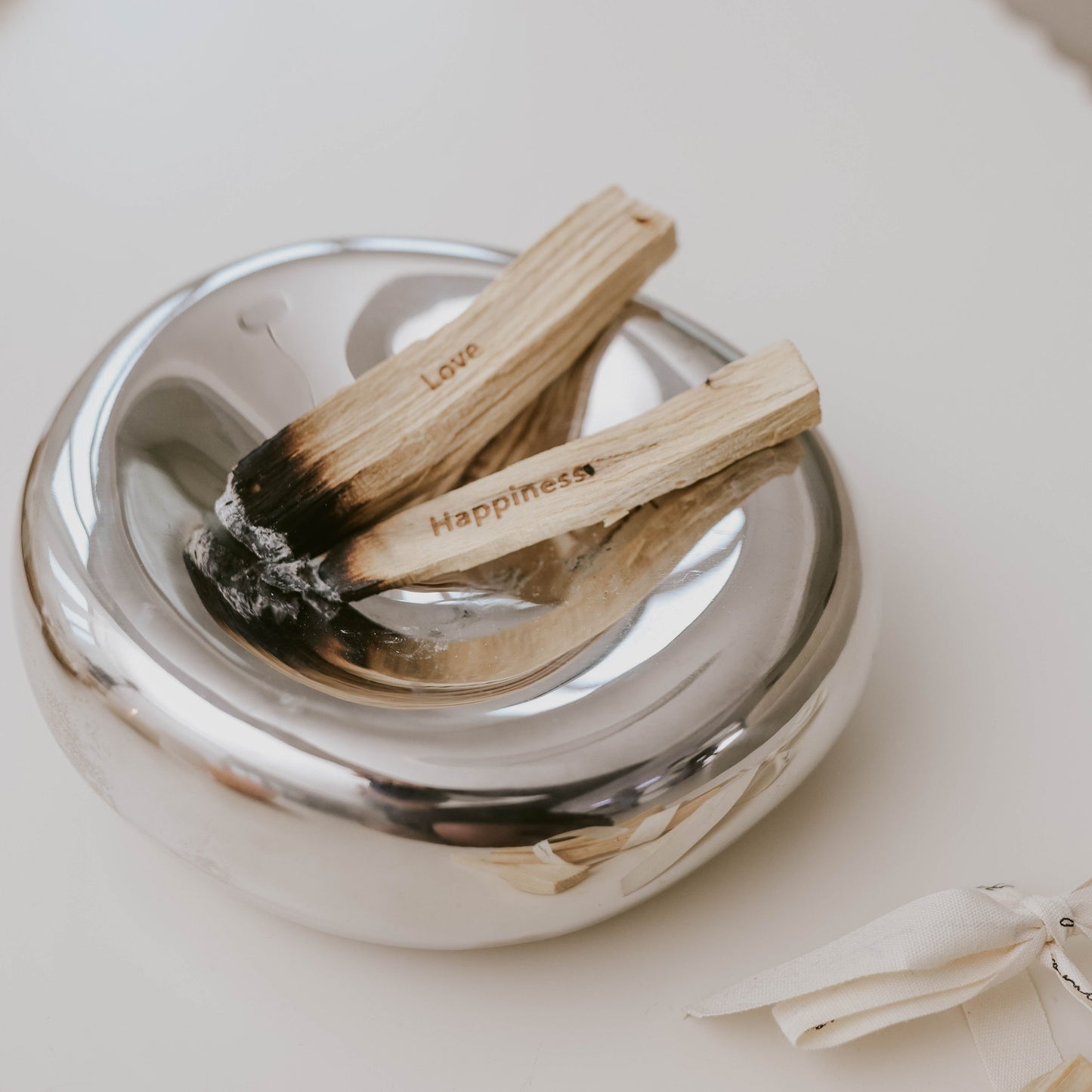 Auf einer feuerfesten spiegelnden, runden Unterlage liegen zwei angezündete Palo Santo mit den Mantras Love und Happiness.