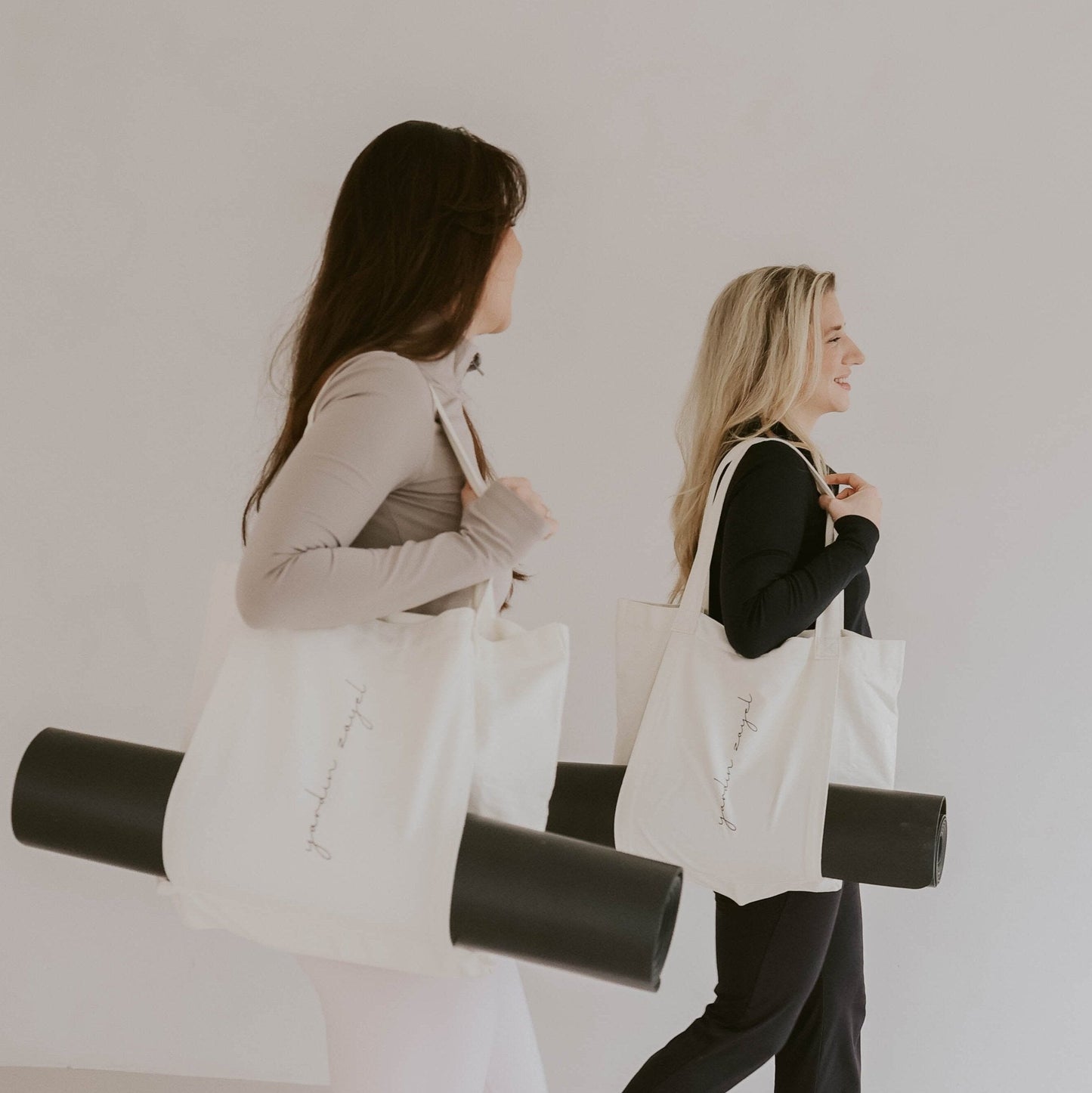 Zwei Freundinnen, blond und dunkelhaarig, sportlich bekleidet, laufen nebeneinander. Beide tragen die Flow Tote Yogatasche mit der DTWW Yogamatte in der Halterung.