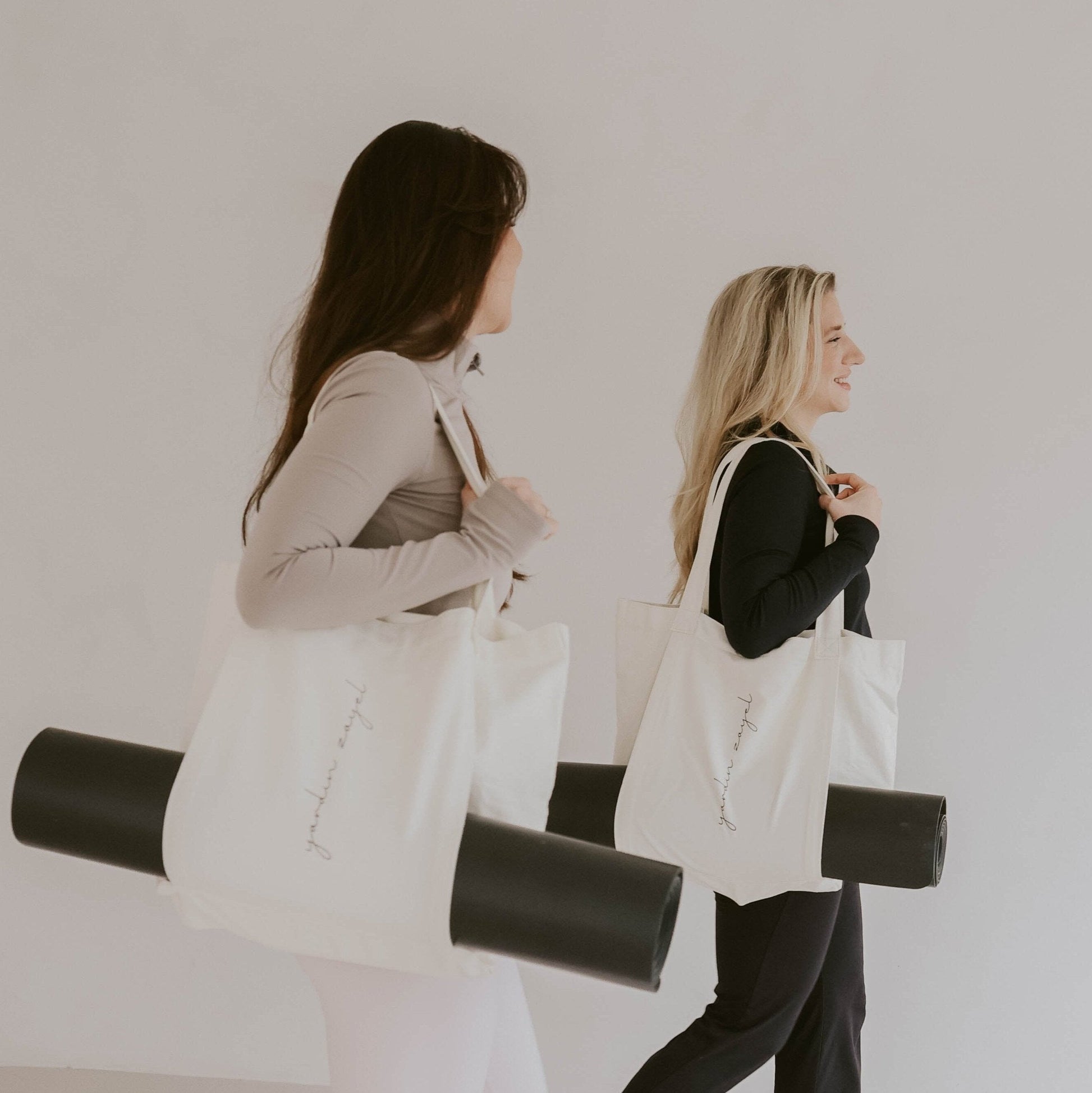 Zwei Freundinnen, blond und dunkelhaarig, sportlich bekleidet, laufen nebeneinander. Beide tragen die Flow Tote Yogatasche mit der DTWW Yogamatte in der Halterung.