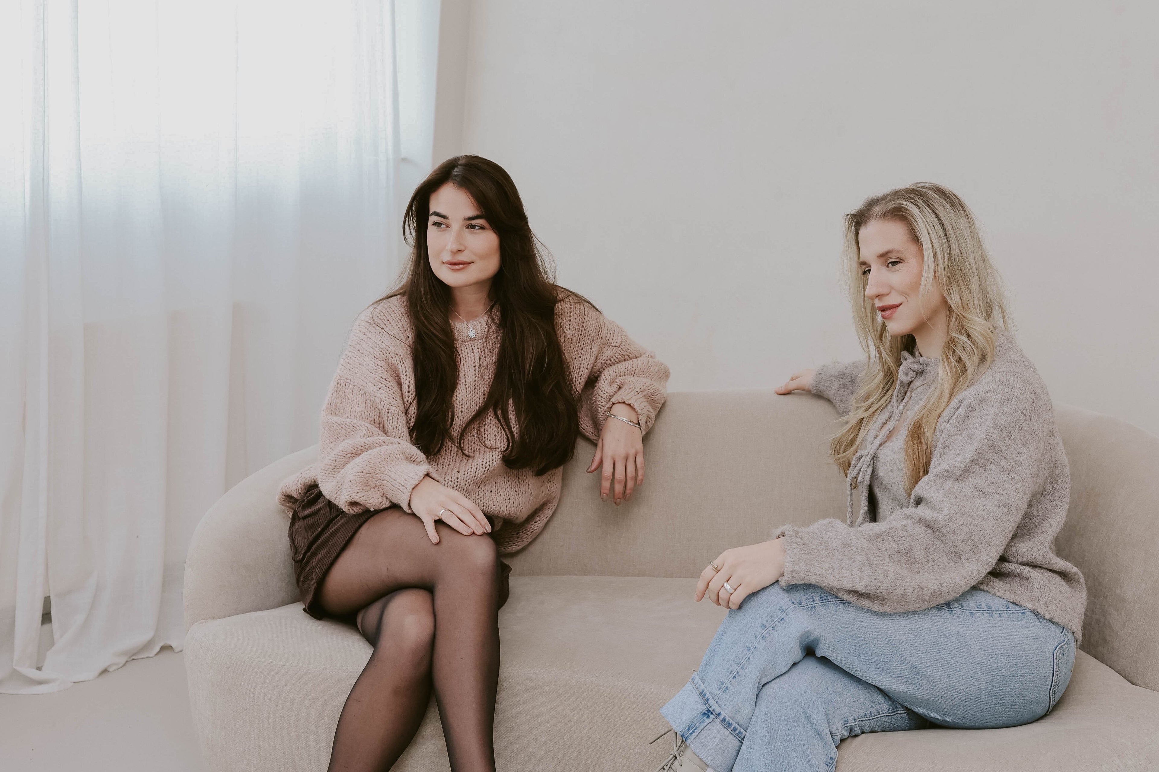Beide Gründerinnen, braunhaarig links in naturtönen gekleidet, rechts, blond in einer Jeans und einem Strickoberteil. Beide sitzen auf einem beigen Sofa und schauen an der Kamera vorbei.
