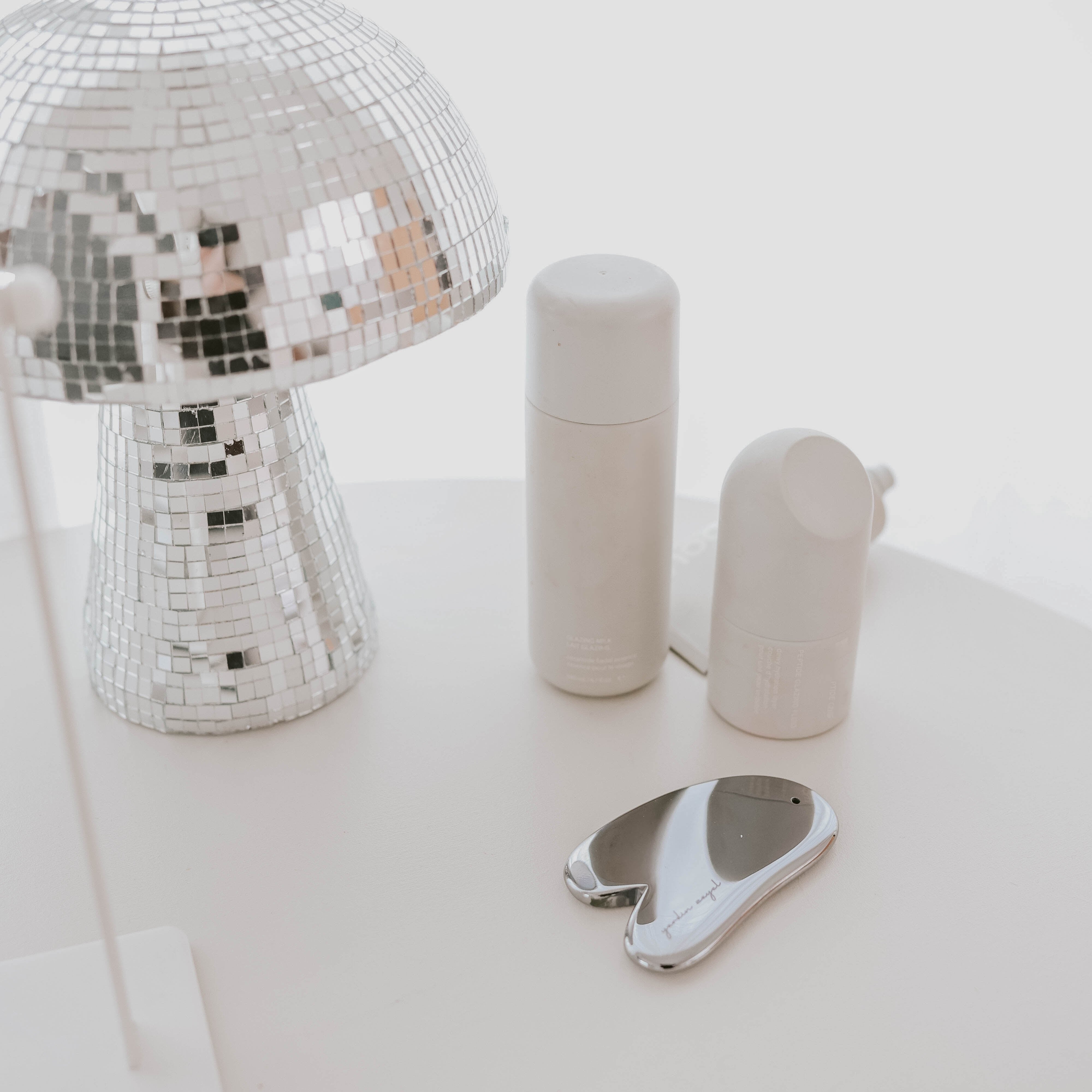 In einer hellen und ruhigen Umgebung ist sind zwei hellgraue Skincare Flaschen, das Teraglow Gua Sha aus Terahertz sowie eine Pilzförmige Diskokugel zu sehen.