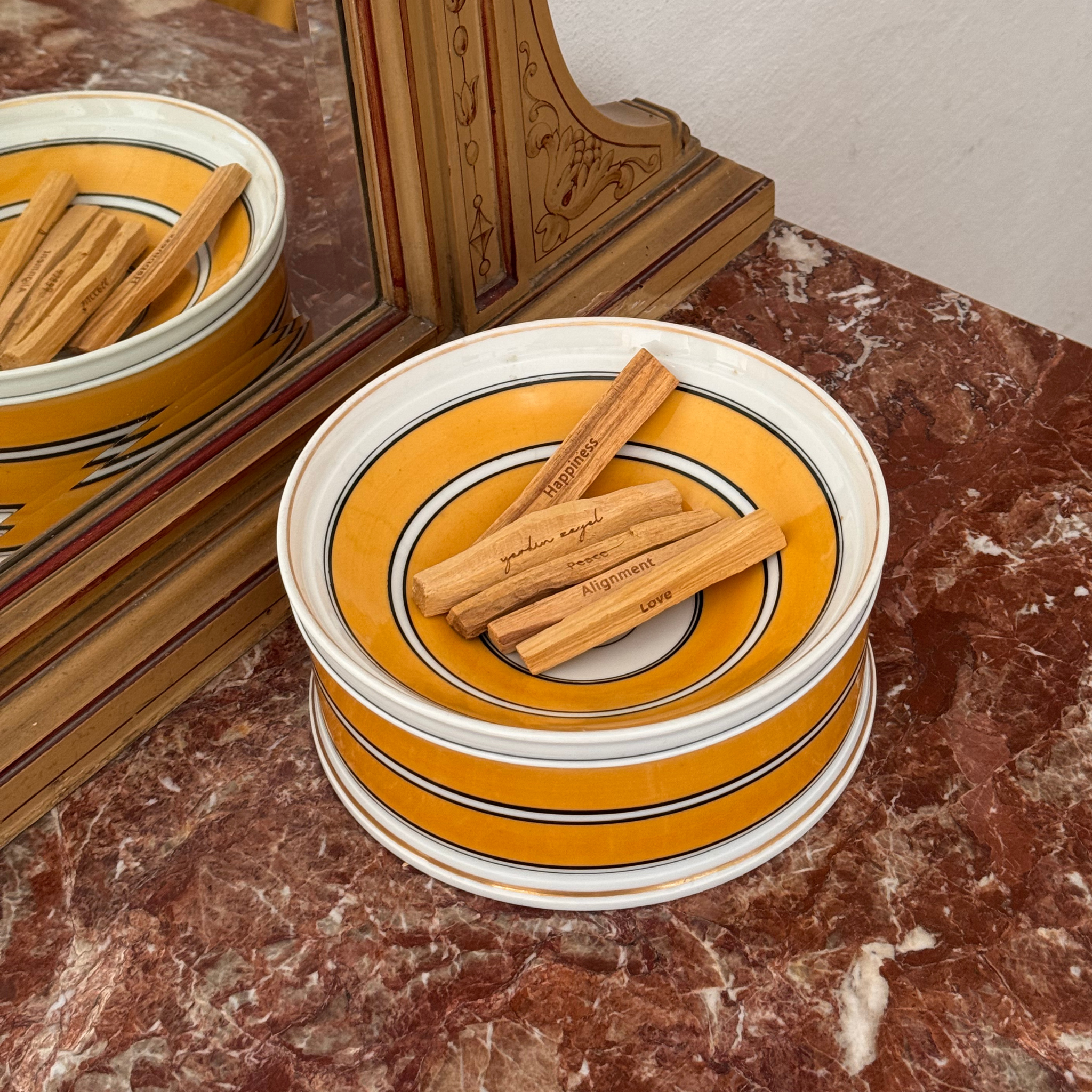 Fünf Palo Santo Zenwood Sticks mit verschiedenen Intentionen auf  einer gelben Schale, stehend auf rotem Marmortisch. Ritual-Setup.